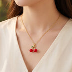 A close-up of a woman wearing a delicate gold chain necklace with a cherry pendant, styled with a cream-colored V-neck top against a soft, blurred outdoor background.