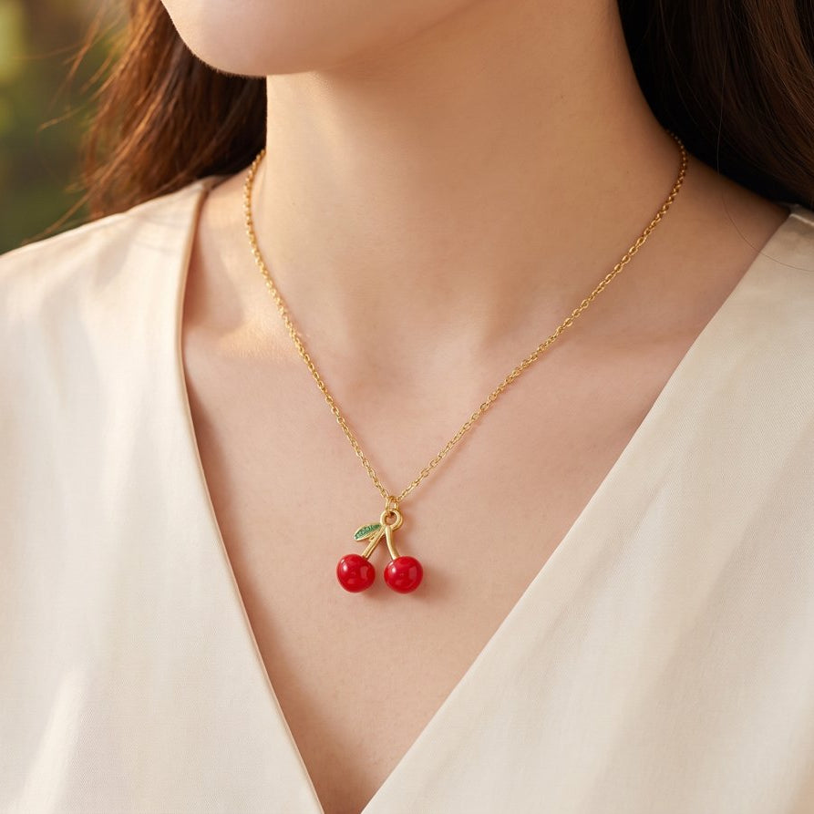 A close-up of a woman wearing a delicate gold chain necklace with a cherry pendant, styled with a cream-colored V-neck top against a soft, blurred outdoor background.